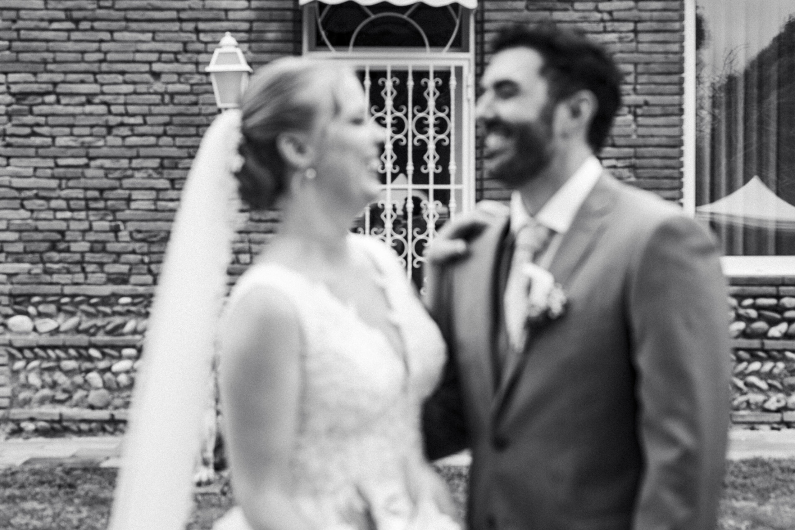 Mariés souriants en noir et blanc devant une bâtisse en briques, capturant un moment de complicité.