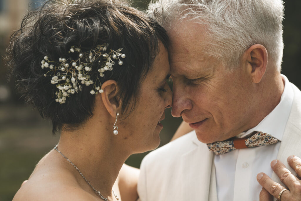 Un couple de mariés se touche tendrement le front en partageant un moment intime. La mariée porte une coiffure ornée de fleurs blanches et de perles, et le marié sourit affectueusement.