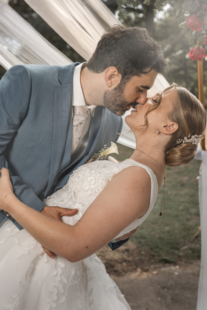 une photographie d'un marié penche sa mariée pour un baiser romantique, capturé en pleine lumière naturelle. Elle porte une robe en dentelle blanche et un chignon orné de perles, tandis que lui est vêtu d’un costume bleu élégant.