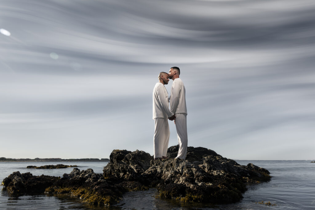 Deux hommes vêtus de blanc, debout sur un rocher au bord de la mer, se tenant la main et échangeant un moment tendre sous un ciel nuageux.