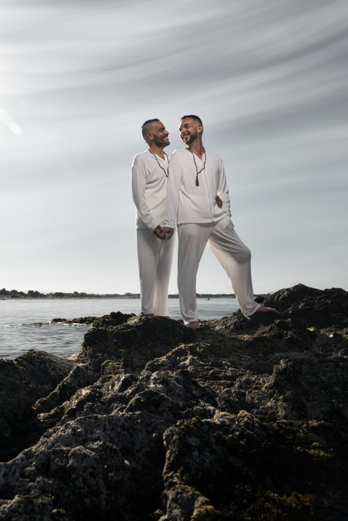 Deux hommes
complices, habillés en blanc, se tenant la main et échangeant un sourire sur des rochers face à la mer.