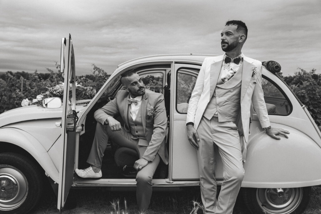 Deux hommes en costume posent élégamment avec une voiture 2CV décorée, dans un vignoble, lors de leur mariage, en noir et blanc.