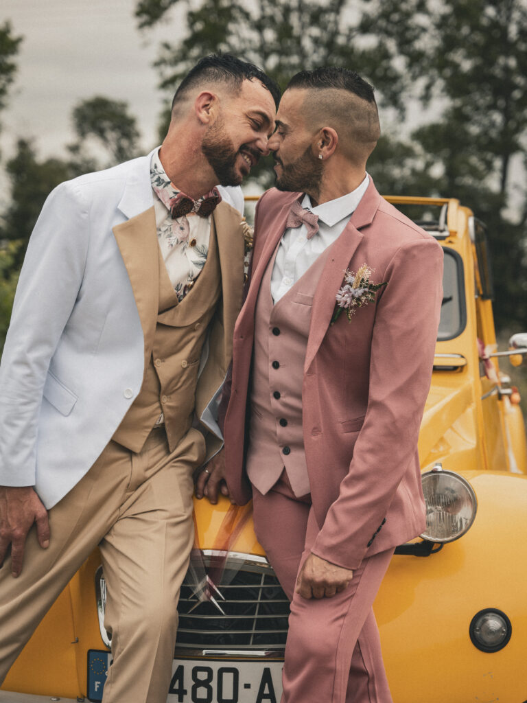 Deux hommes en costume pastel se touchant tendrement le front devant une voiture ancienne jaune, symbolisant leur union et leur complicité.