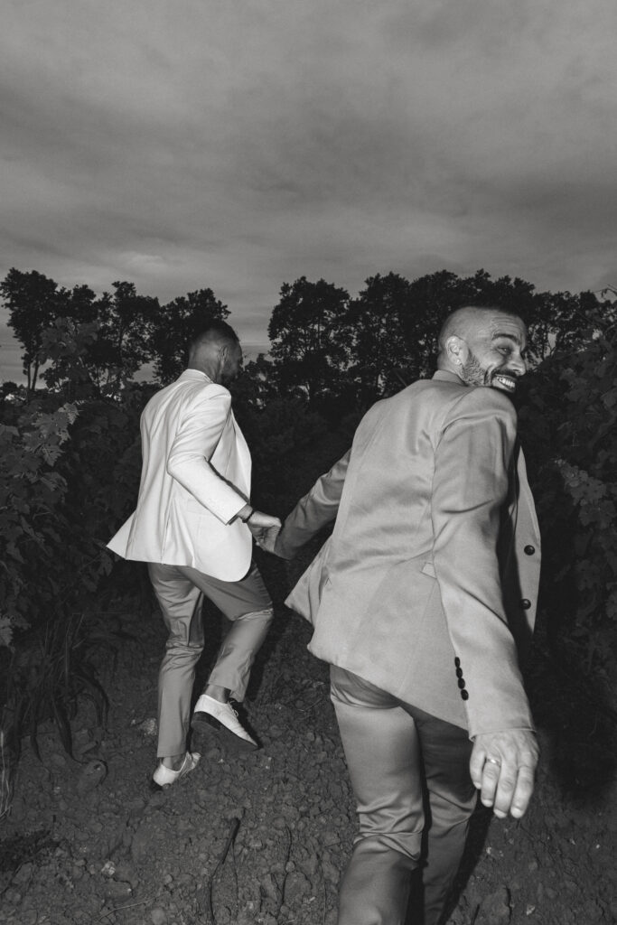 Deux hommes élégamment habillés en costume se tiennent la main en marchant dans un chemin de vigne, regard complice et sourire, capturés en noir et blanc.