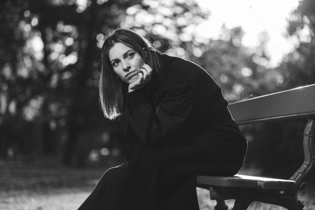 Portrait en noir et blanc d'une femme pensive sur un banc, capturé en contre-jour. Un cliché artistique jouant sur la lumière et l'émotion.