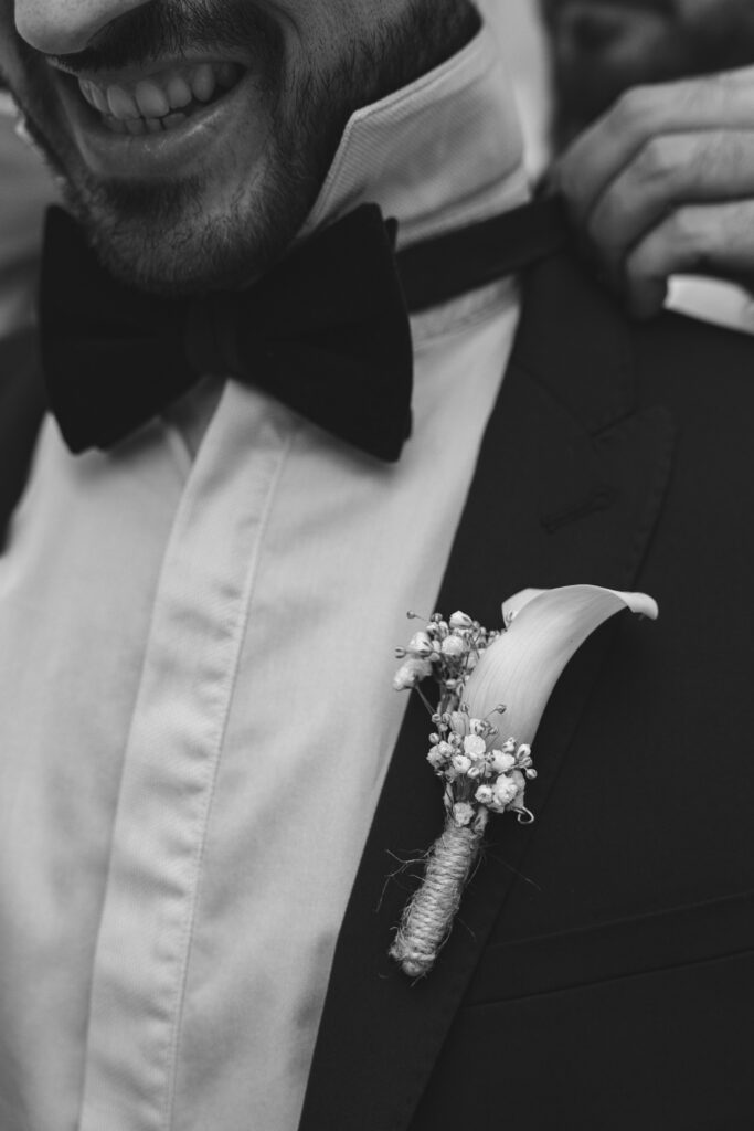 photographie en noir et blanc d'un invité de mariage se faisant mettre un nœud papillon avant la cérémonie.