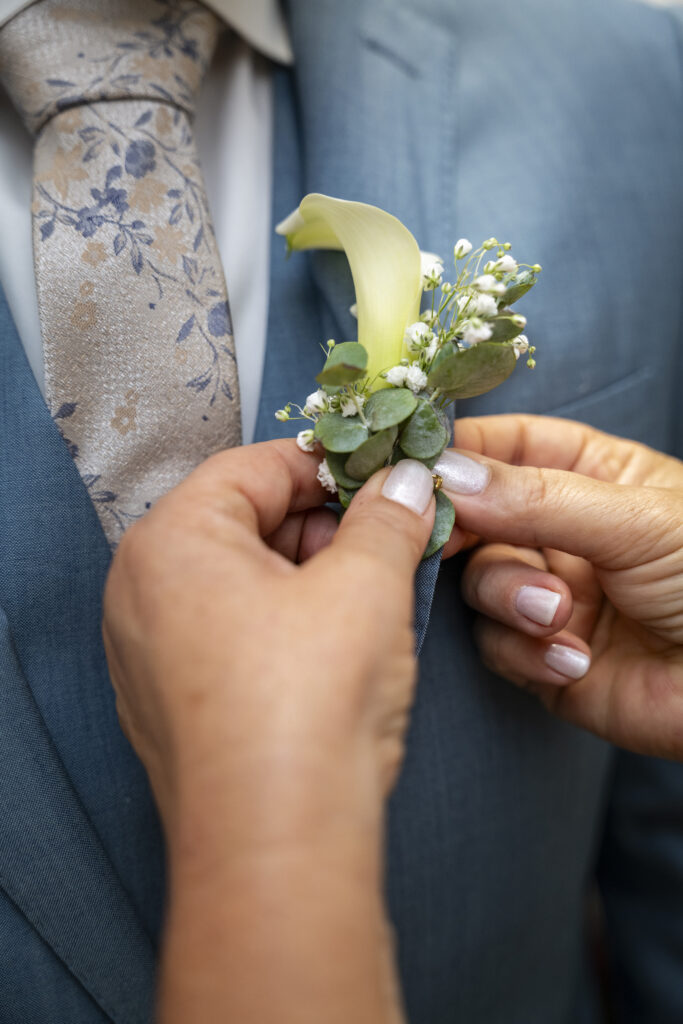 photographie d'une main attachant une boutonnière élégante à un costume bleu marine à un mariage.