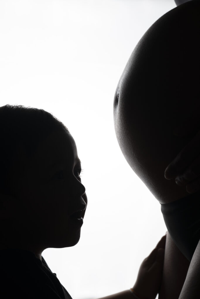 Portrait artistique en contre-jour d’un enfant touchant le ventre arrondi de sa mère enceinte – photo de maternité intime et émotionnelle réalisée par un photographe professionnel.