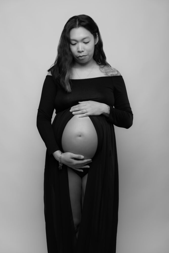 Portrait en studio d’une femme enceinte en robe noire, les mains posées sur son ventre arrondi – photo de grossesse élégante en noir et blanc.